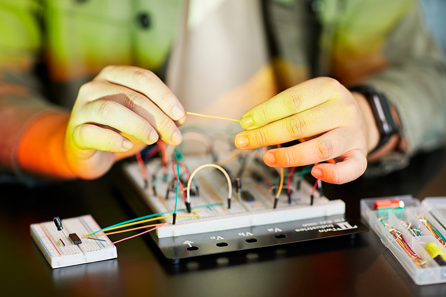 Image shows an engineering student sitting at a table at home working on a circuit board included in a at-home lab kit.