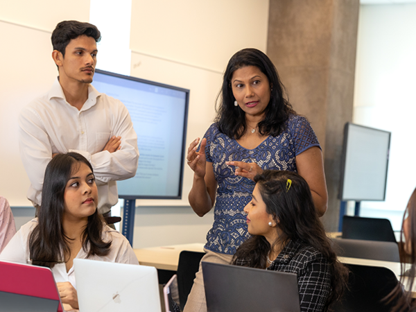 A group of people engaged in discussion in a conference room, with laptops and a screen in the background.